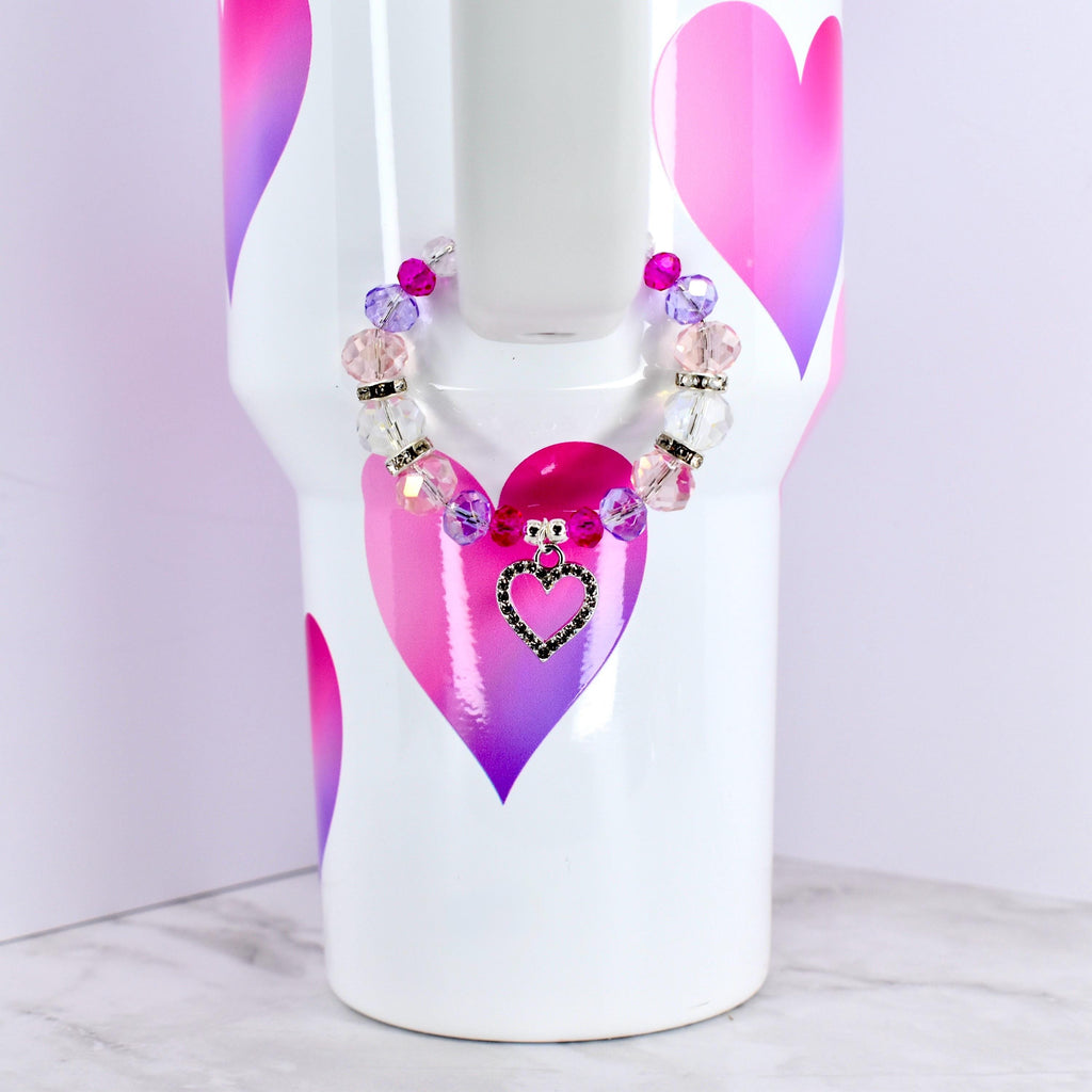 A white tumbler with pink and purple heart designs, adorned with a beaded bracelet. The bracelet features clear, pink, and purple beads, and a heart-shaped charm with a black outline and a black center.