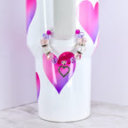 A white tumbler with pink and purple heart designs, adorned with a beaded bracelet. The bracelet features clear, pink, and purple beads, and a heart-shaped charm with a black outline and a black center.