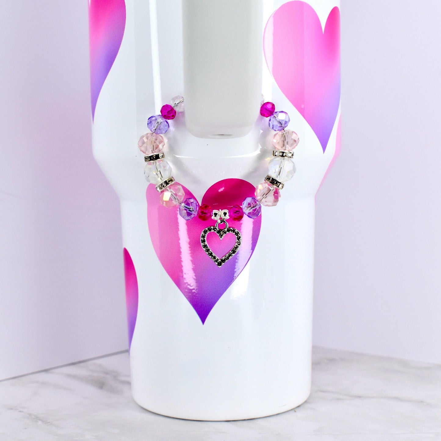 A white tumbler with pink and purple heart designs, adorned with a beaded bracelet. The bracelet features clear, pink, and purple beads, and a heart-shaped charm with a black outline and a black center.