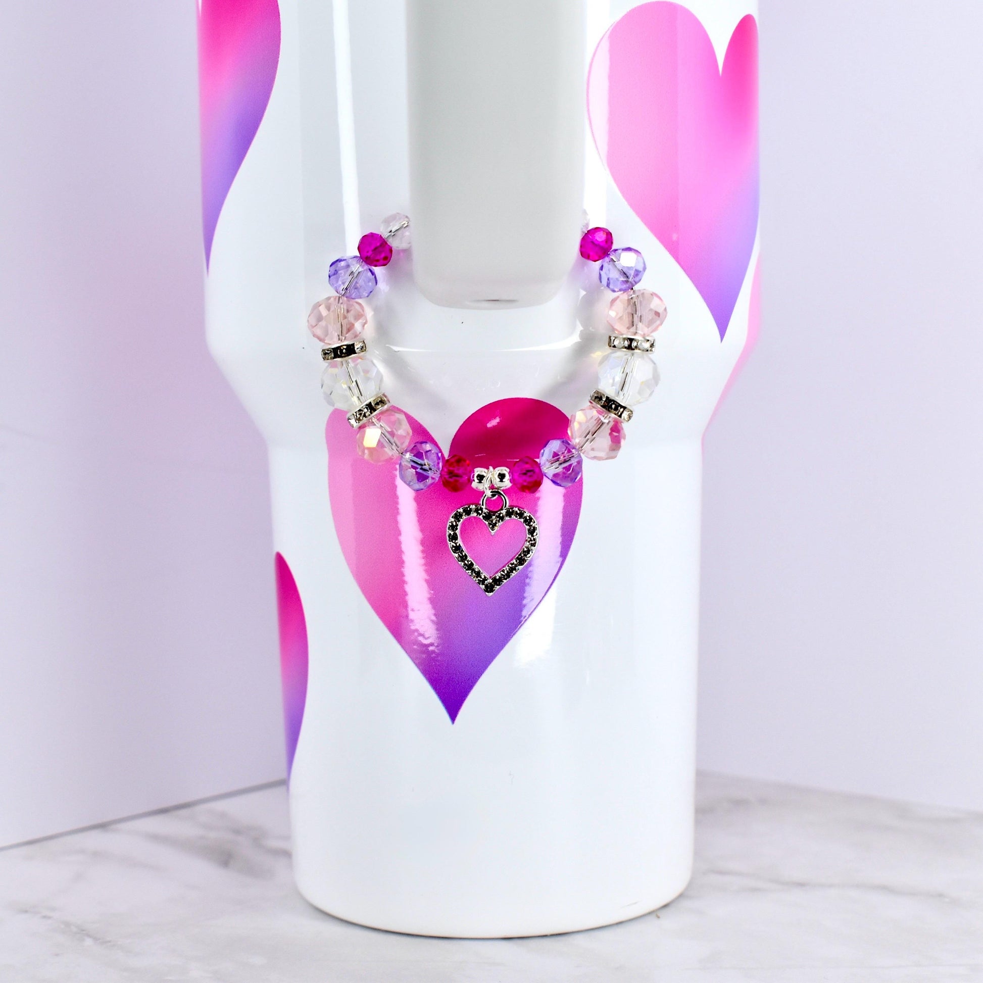 A white tumbler with pink and purple heart designs, adorned with a beaded bracelet. The bracelet features clear, pink, and purple beads, and a heart-shaped charm with a black outline and a black center.