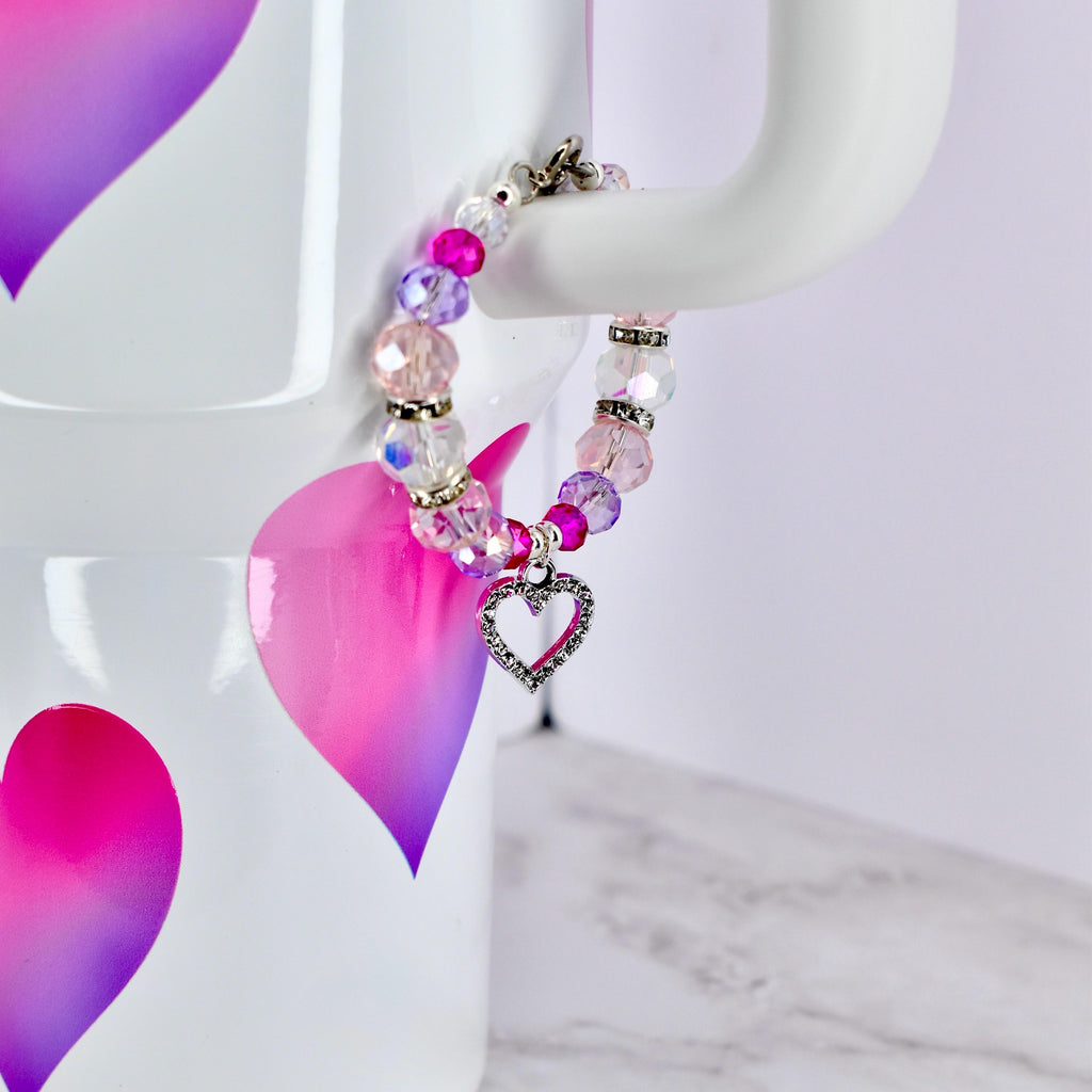 A beaded bracelet with a heart charm. The bracelet features a mix of pink, purple, and clear beads, with a silver-colored heart charm. The beads are strung together with a silver-colored clasp. The bracelet is displayed against a white background.