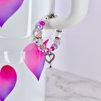 A beaded bracelet with a heart charm. The bracelet features a mix of pink, purple, and clear beads, with a silver-colored heart charm. The beads are strung together with a silver-colored clasp. The bracelet is displayed against a white background.