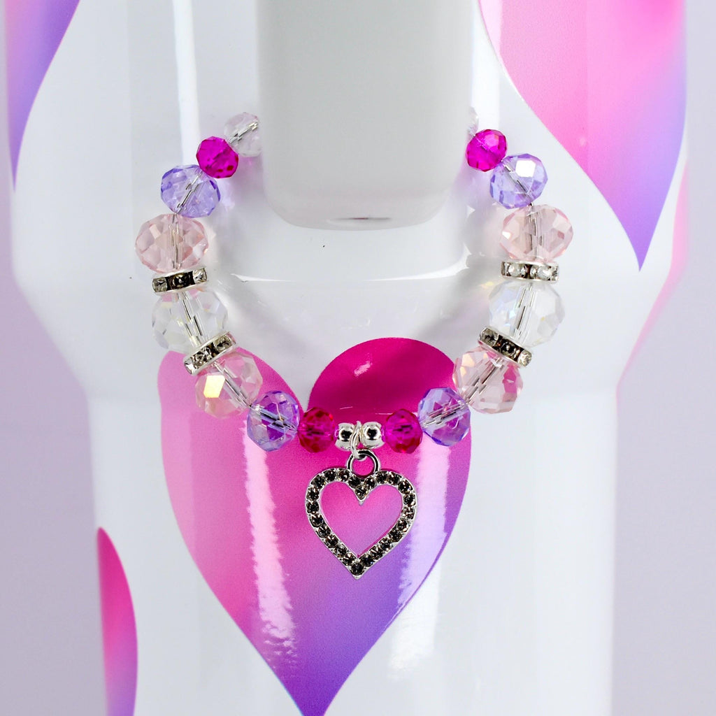 A beaded bracelet with pink, purple, and clear beads, and a silver heart charm. The bracelet is displayed on a white surface with a pink and purple heart design.