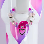 A beaded bracelet with pink, purple, and clear beads, and a silver heart charm. The bracelet is displayed on a white surface with a pink and purple heart design.