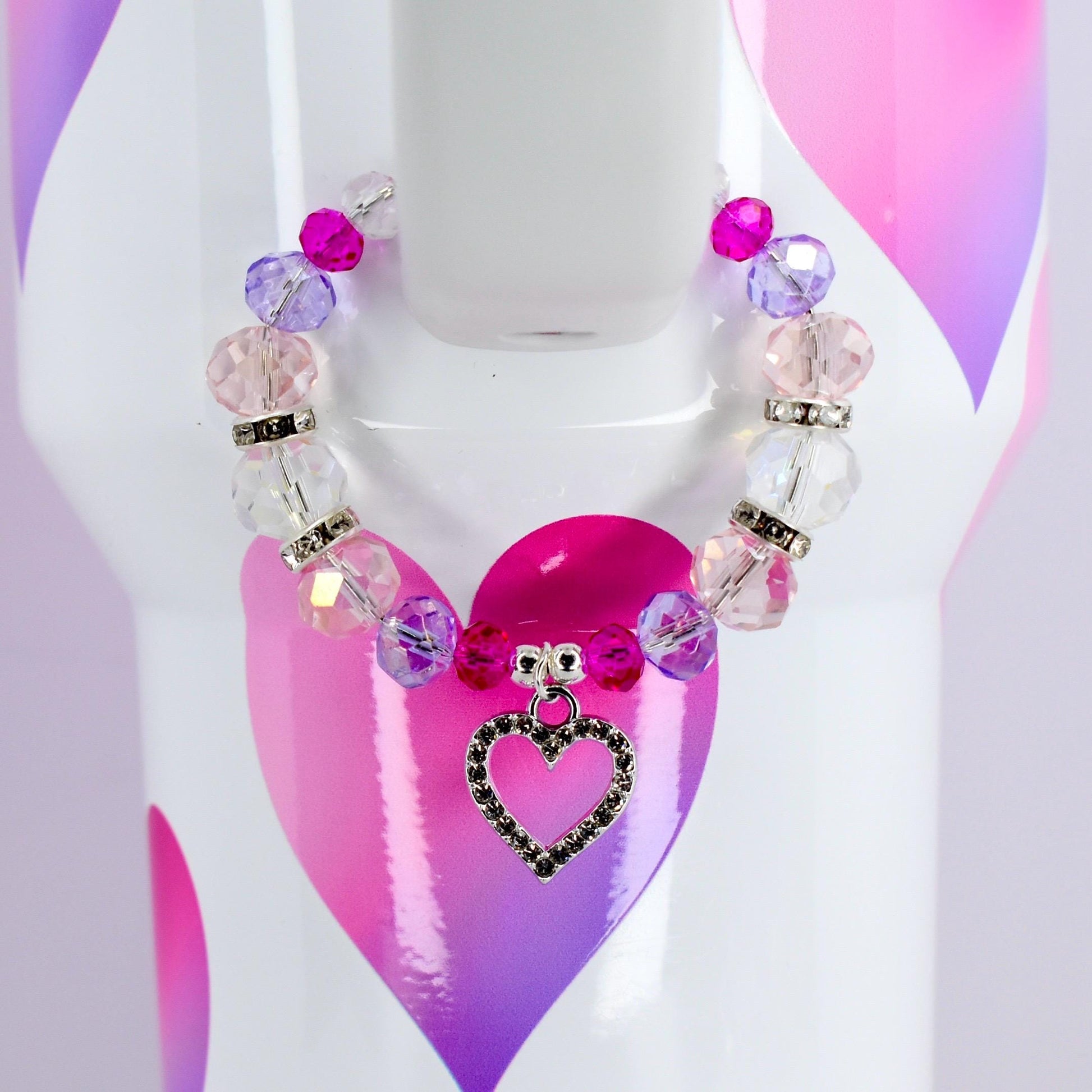 A beaded bracelet with pink, purple, and clear beads, and a silver heart charm. The bracelet is displayed on a white surface with a pink and purple heart design.