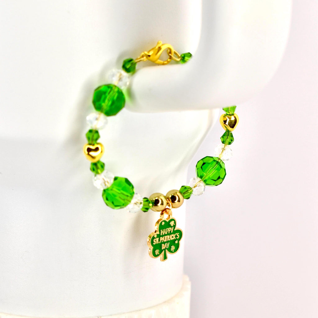 A Saint Patrick's Day bracelet featuring green and clear beads, gold accents, and a charm with a shamrock and the text "Happy St. Patrick's Day". The bracelet is designed with a gold clasp.