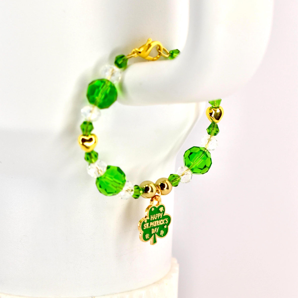 A Saint Patrick's Day bracelet featuring green and clear beads, gold accents, and a charm with a shamrock and the text "Happy St. Patrick's Day". The bracelet is designed with a gold clasp.