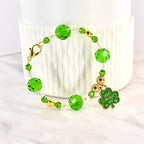 A beaded bracelet with green and clear beads, gold accents, and a gold clasp. A green shamrock charm with "Happy St. Patrick's Day" text hangs from the bracelet. The bracelet is displayed on a white surface.