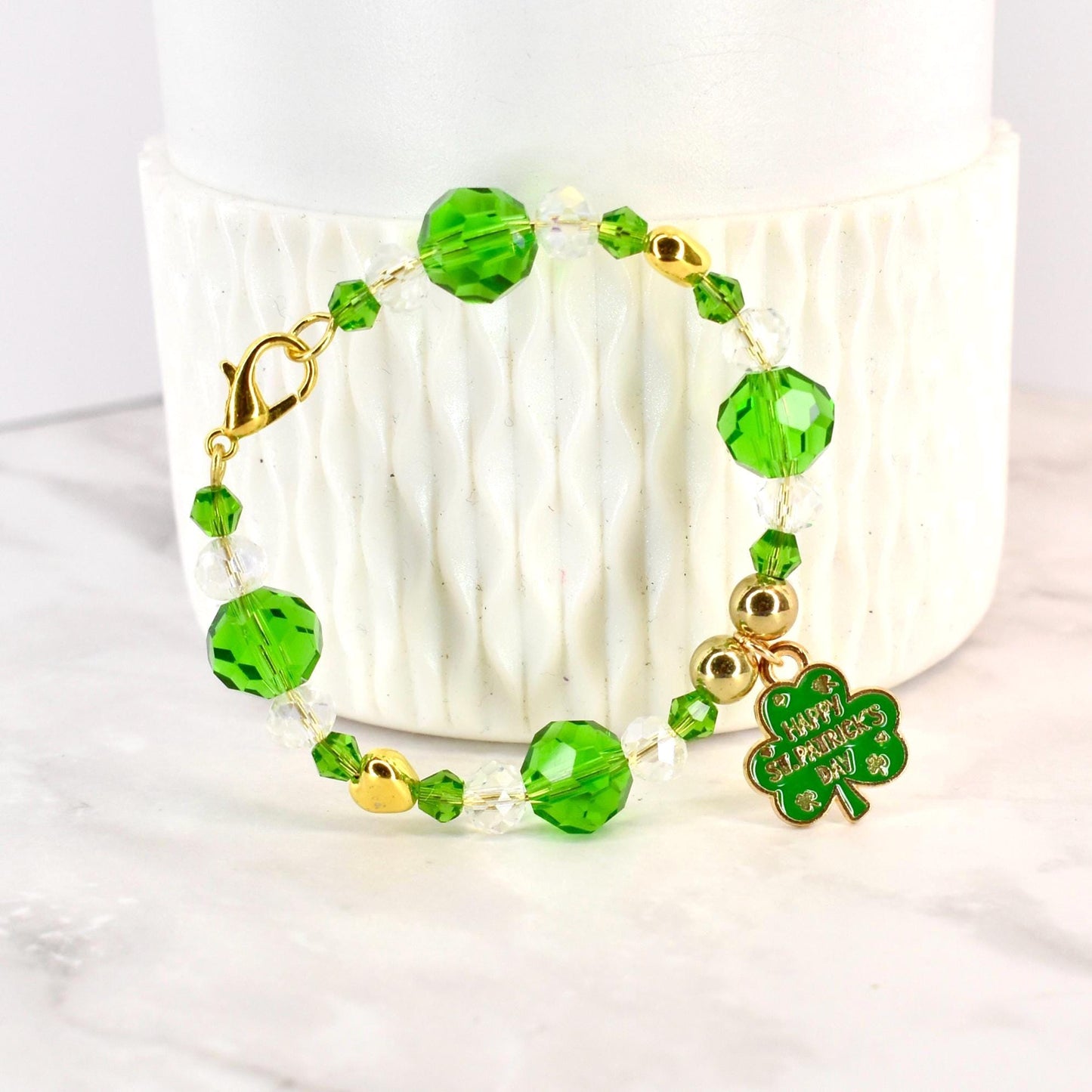A beaded bracelet with green and clear beads, gold accents, and a gold clasp. A green shamrock charm with "Happy St. Patrick's Day" text hangs from the bracelet. The bracelet is displayed on a white surface.