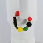 A gold-toned handle bracelet featuring a vibrant design. The bracelet is adorned with a series of colorful beads in red, black, yellow, and green, accented with gold-colored spacers. A small, stylized shape with red, black, and green colors is also present.
