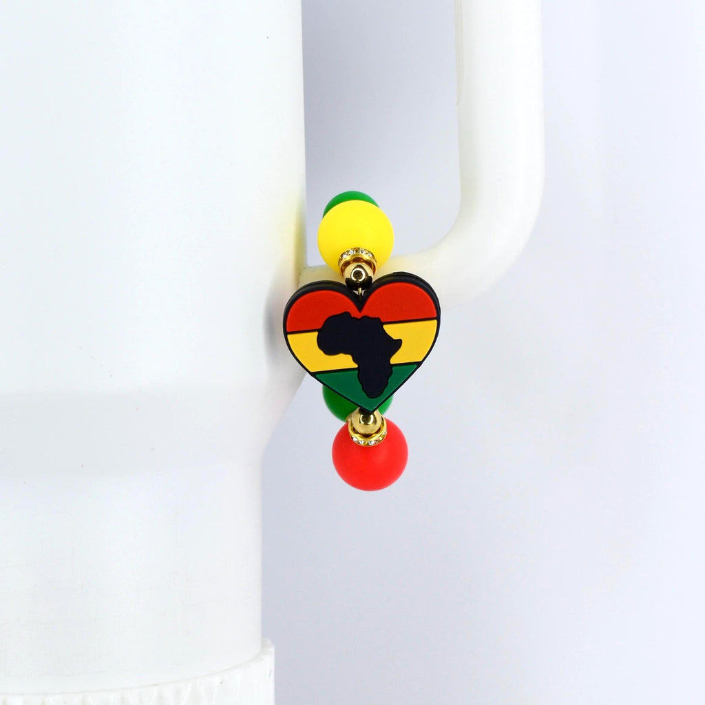 A colorful handle bracelet featuring a heart-shaped pendant with the outline of Africa in black. The heart is colored in red, yellow, and green, with yellow and green beads above and a red bead below. The bracelet is attached to a white object.