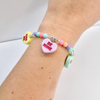 A beaded bracelet with pastel-colored beads and heart-shaped charms. The charms read "KISS ME" and "BE MINE" in red text. The bracelet is a mix of colors, including yellow, pink, green, and purple.