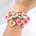 A stack of colorful beaded bracelets with heart-shaped charms. The charms have phrases like "Kiss Me," "Sweet Heart," and "True Love" in red lettering. The beads are pastel colors and clear. A fun, romantic accessory.