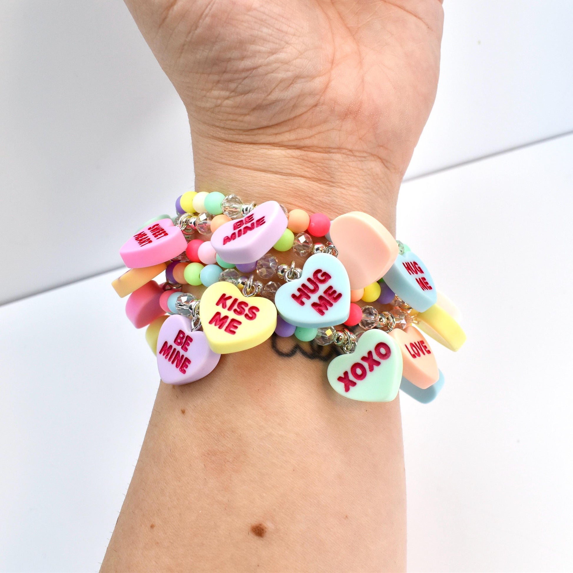 A multi-strand bracelet featuring pastel-colored beads and heart-shaped charms. The charms display phrases like "Kiss Me," "Be Mine," and "XOXO" in red lettering. The bracelet is worn on a wrist.