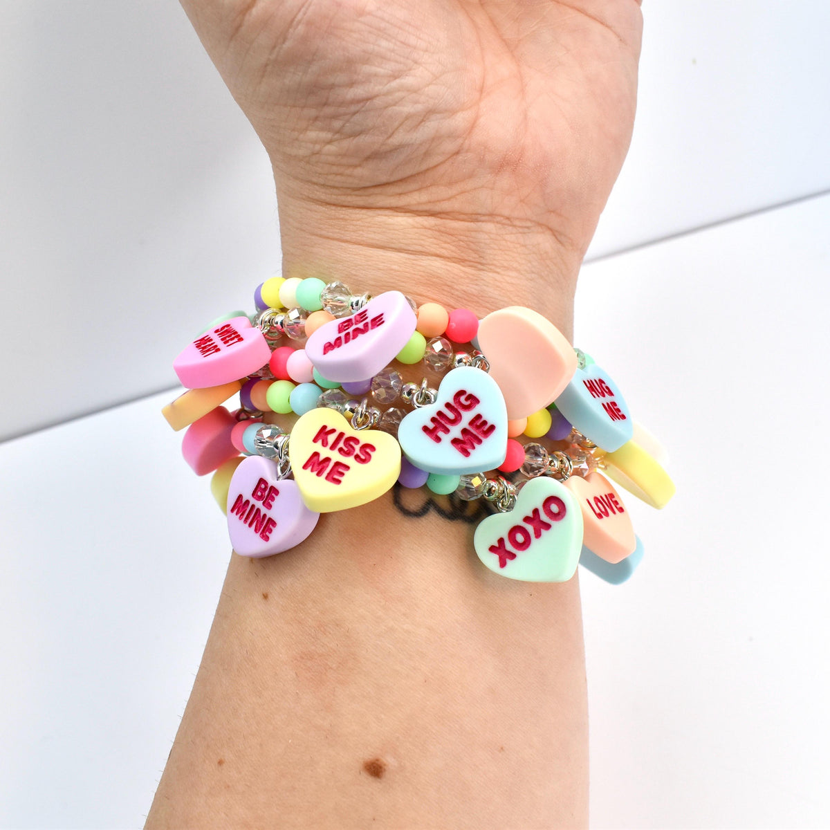 A multi-strand bracelet featuring pastel-colored beads and heart-shaped charms. The charms display phrases like "Kiss Me," "Be Mine," and "XOXO" in red lettering. The bracelet is worn on a wrist.