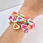 A stack of colorful beaded bracelets with heart-shaped charms. The charms are pastel colors and have phrases like "Kiss Me", "Be Mine", and "Sweet Heart". The bracelets are on a wrist.