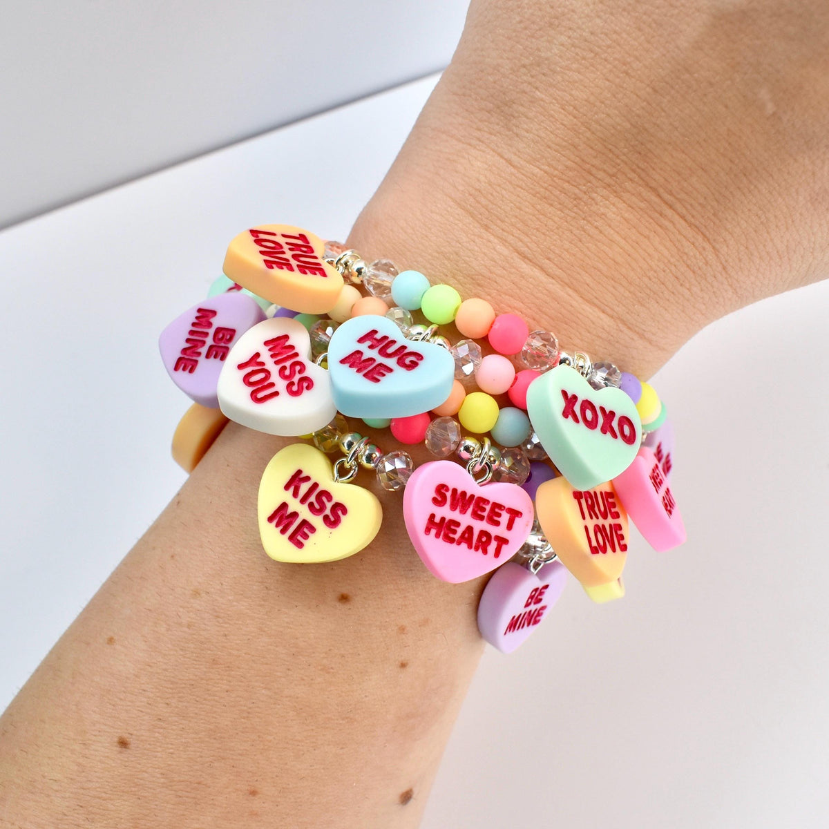 A stack of colorful beaded bracelets with heart-shaped charms. The charms are pastel colors and have phrases like "Kiss Me", "Be Mine", and "Sweet Heart". The bracelets are on a wrist.