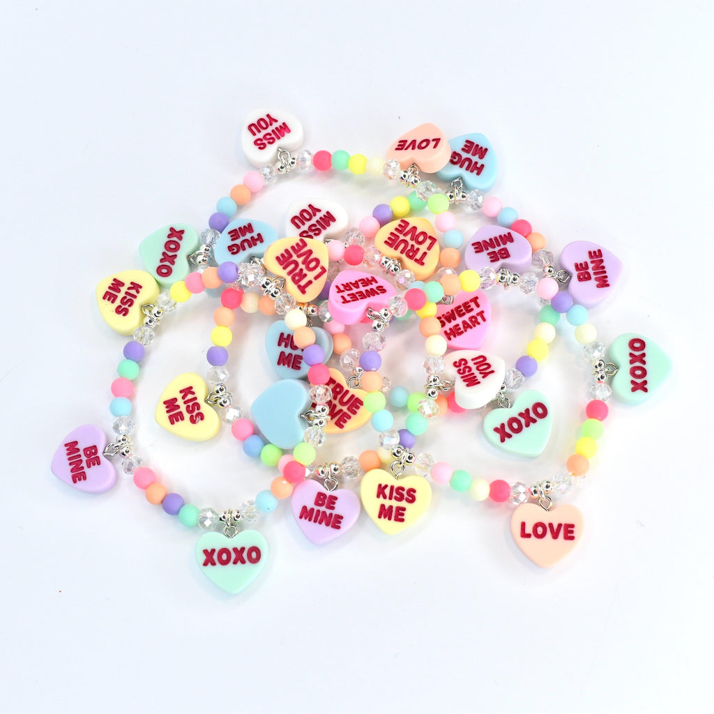 A collection of colorful beaded bracelets with heart-shaped pendants. Each pendant features a different romantic message in red text, such as "Kiss Me," "Be Mine," and "Love." The beads are pastel colors, and the necklaces have silver-colored clasps.