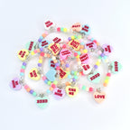 A collection of colorful beaded bracelets with heart-shaped pendants. Each pendant features a different romantic message in red text, such as "Kiss Me," "Be Mine," and "Love." The beads are pastel colors, and the necklaces have silver-colored clasps.
