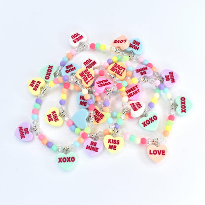 A collection of colorful beaded bracelets with heart-shaped pendants. Each pendant features a different romantic message in red text, such as "Kiss Me," "Be Mine," and "Love." The beads are pastel colors, and the necklaces have silver-colored clasps.
