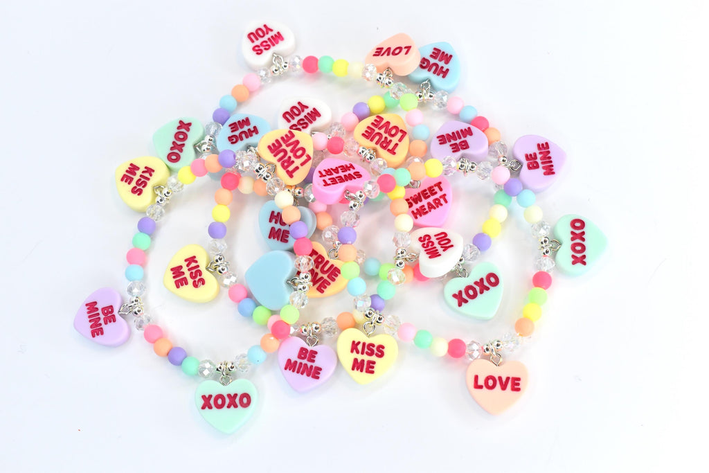 A collection of colorful beaded bracelets with heart-shaped charms. The charms feature phrases like "Kiss Me," "Be Mine," and "XOXO" in red text. The beads are pastel colors, and the bracelets have silver-colored accents.