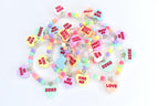 A collection of colorful beaded bracelets with heart-shaped charms. The charms feature phrases like "Kiss Me," "Be Mine," and "XOXO" in red text. The beads are pastel colors, and the bracelets have silver-colored accents.