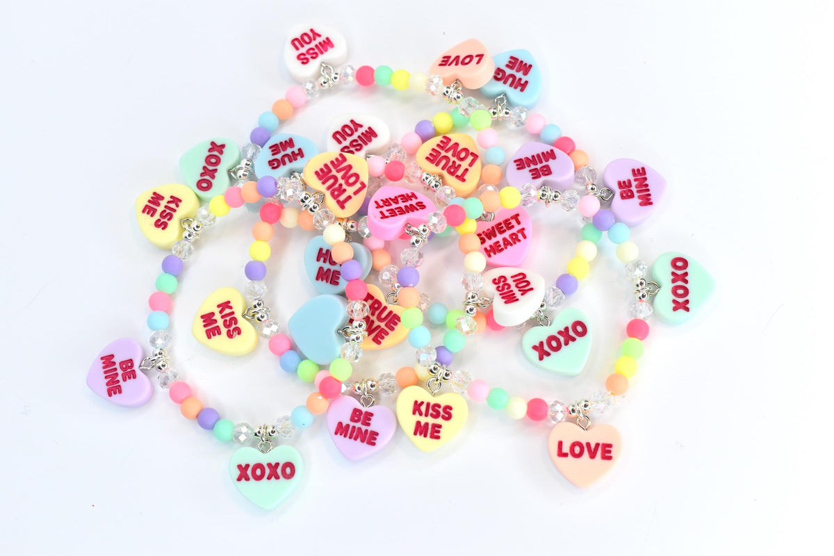 A collection of colorful beaded bracelets with heart-shaped charms. The charms feature phrases like "Kiss Me," "Be Mine," and "XOXO" in red text. The beads are pastel colors, and the bracelets have silver-colored accents.