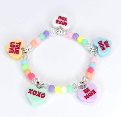 A colorful beaded bracelet with heart-shaped charms. The charms have messages like "TRUE LOVE," "MISS YOU," "HUG ME," "BE MINE," and "XOXO." The beads are pastel colors, and the bracelet has silver-colored accents.