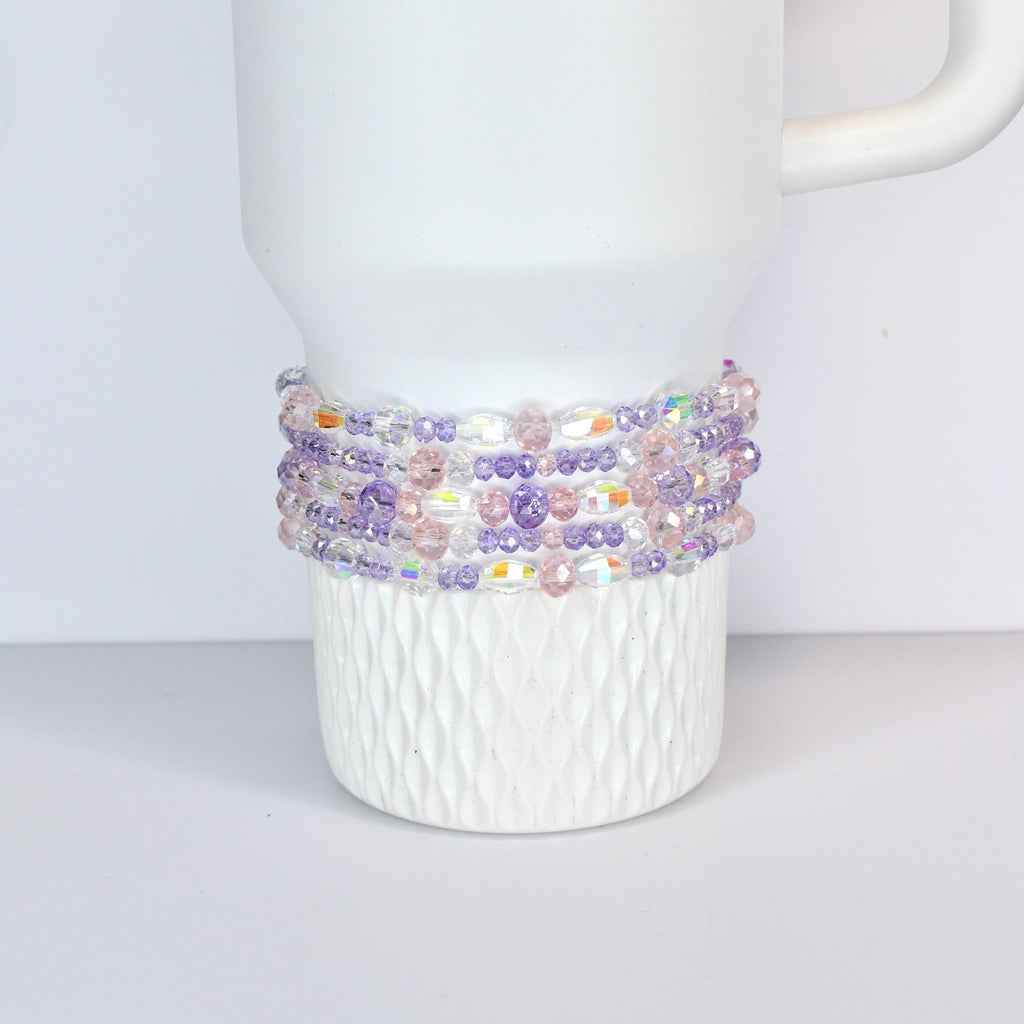 A white Stanley with a beaded wrap. The wrap features a mix of clear, iridescent, pink, and purple beads. The beads are strung together to create a decorative band around the mug, adding a touch of color and texture.