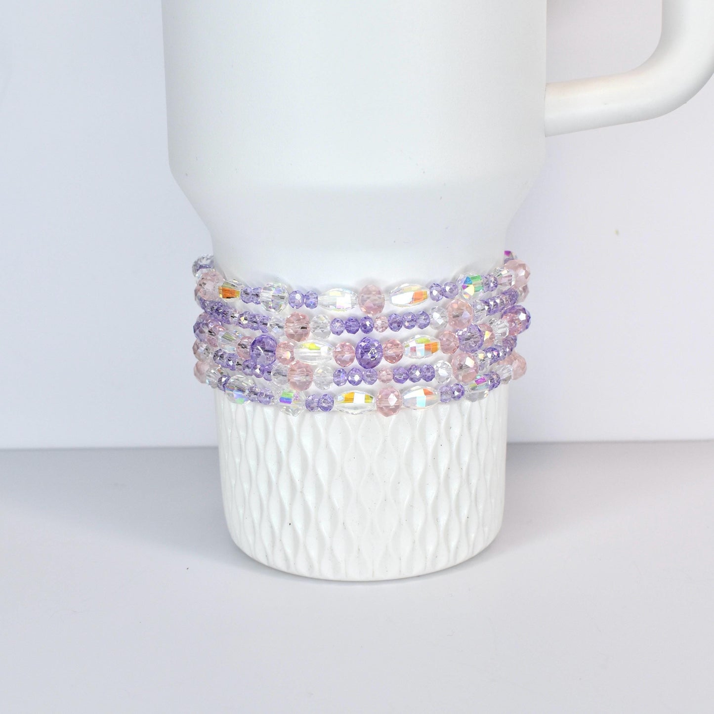 A white Stanley with a beaded wrap. The wrap features a mix of clear, iridescent, pink, and purple beads. The beads are strung together to create a decorative band around the mug, adding a touch of color and texture.