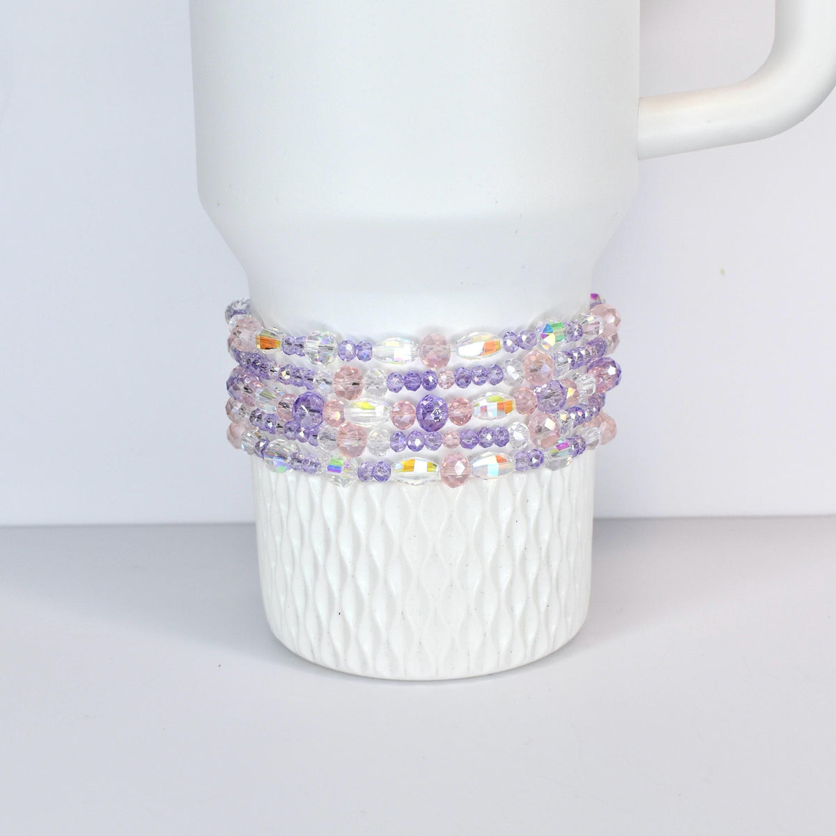 A white Stanley with a beaded wrap. The wrap features a mix of clear, iridescent, pink, and purple beads. The beads are strung together to create a decorative band around the mug, adding a touch of color and texture.