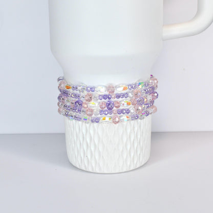 A white Stanley with a beaded wrap. The wrap features a mix of clear, iridescent, pink, and purple beads. The beads are strung together to create a decorative band around the mug, adding a touch of color and texture.