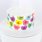 A white silicone tumbler boot decorated with colorful candy heart graphics. The hearts feature phrases like "Be Mine," "Kiss Me," and "Soul Mate." The tumbler boot is on a white surface.