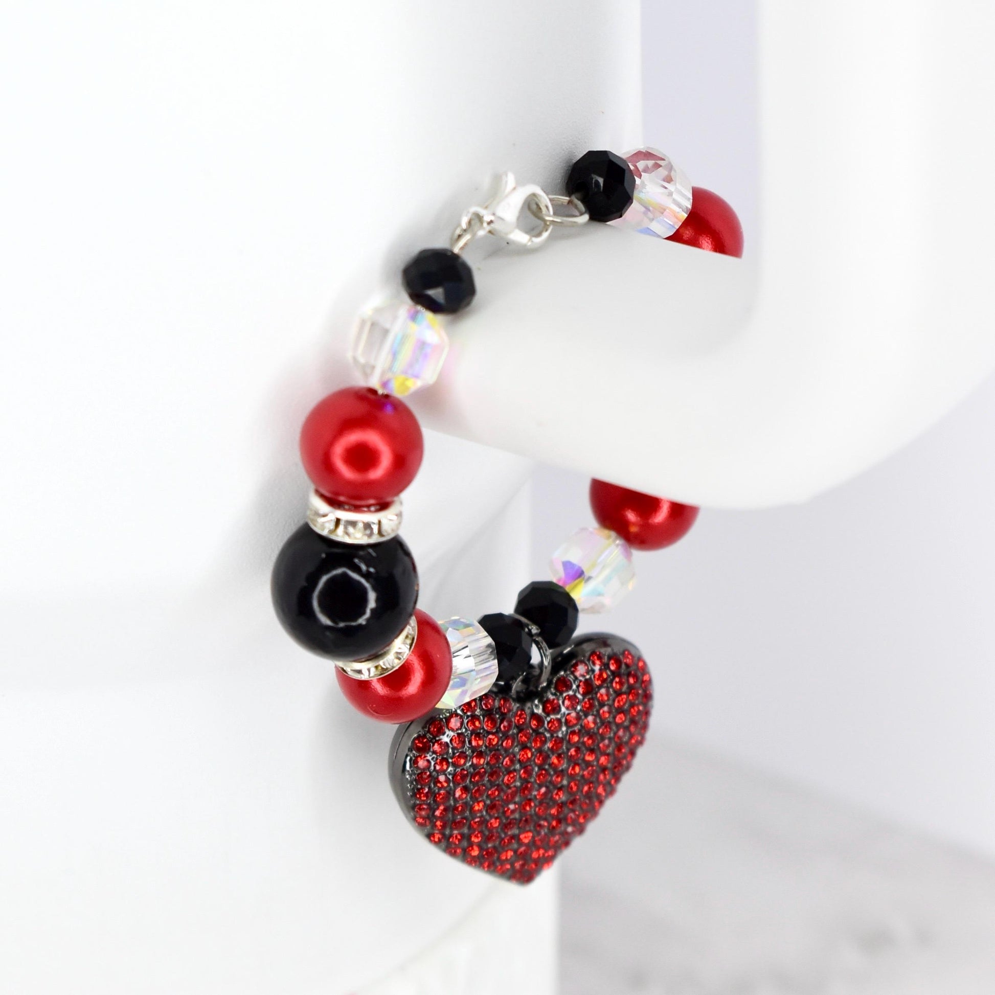 A beaded tumbler bracelet featuring red and black beads, clear iridescent beads, and a red heart-shaped charm covered in red rhinestones. The bracelet has a silver clasp.