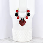A beaded cup charm featuring red, black, and clear beads. A red heart-shaped charm with a rhinestone design hangs from the beads. The charm is designed to fit around a cup or tumbler.
