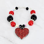 A beaded tumbler bracelet featuring red and black beads, interspersed with clear, iridescent crystals. A heart-shaped pendant, covered in red rhinestones, hangs from the bracelet. The bracelet is connected with a silver clasp.