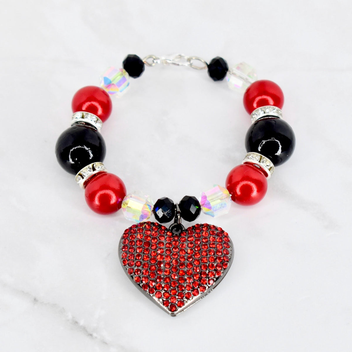 A beaded tumbler bracelet featuring red and black beads, interspersed with clear, iridescent crystals. A heart-shaped pendant, covered in red rhinestones, hangs from the bracelet. The bracelet is connected with a silver clasp.