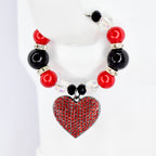 A beaded Stanley handle bracelet featuring red and black beads, clear crystal beads, and a heart-shaped pendant. The heart pendant is covered in small red rhinestones. The bracelet is displayed on a white surface.