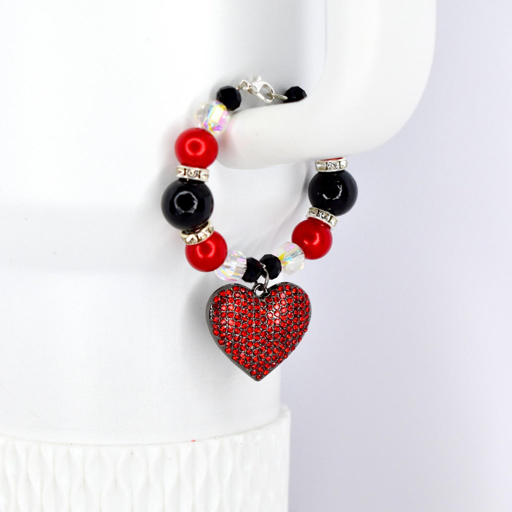 A beaded tumbler handle bracelet featuring red, black, and clear beads. A heart-shaped pendant covered in red rhinestones hangs from the bracelet. The bracelet is held up by a white surface.