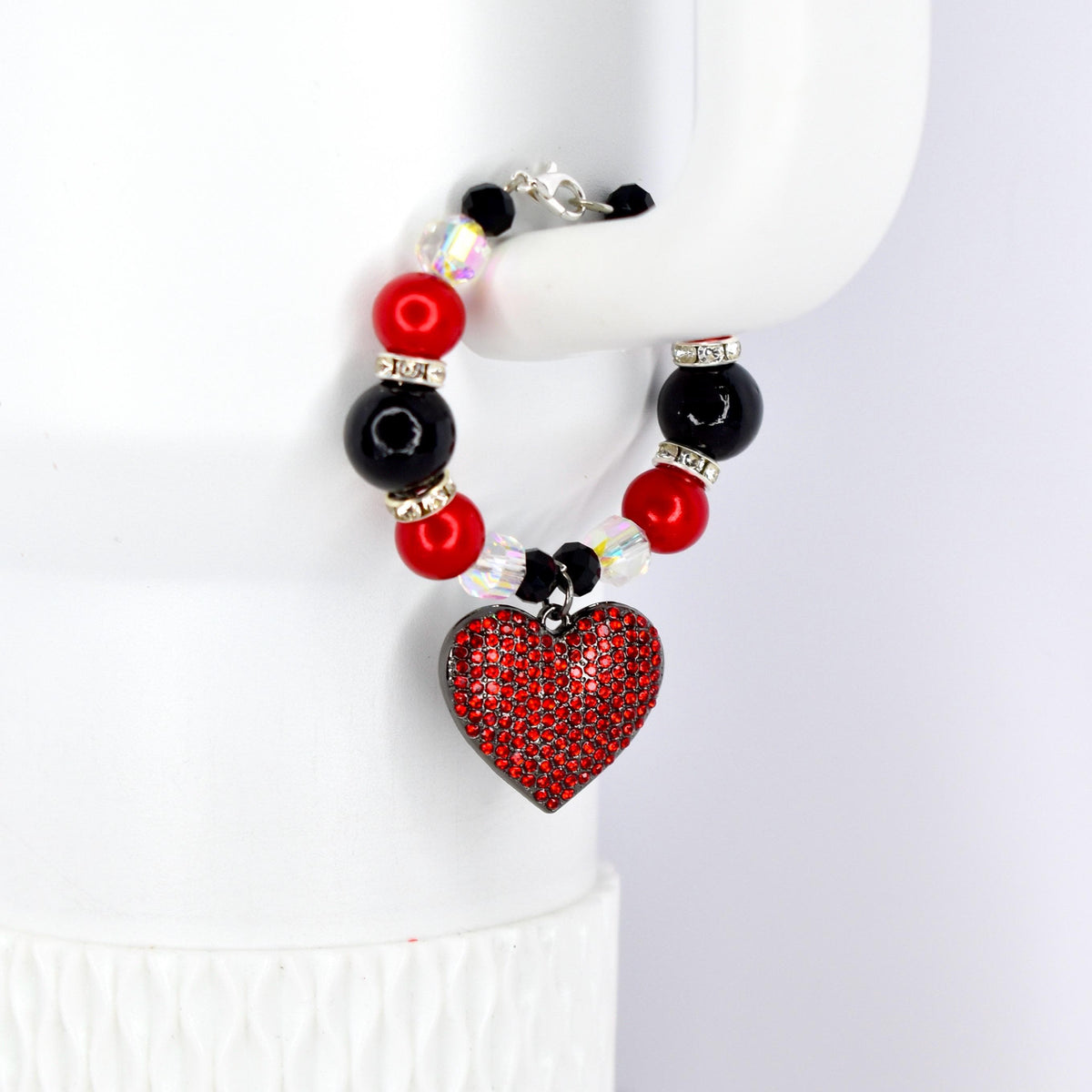 A beaded tumbler handle bracelet featuring red, black, and clear beads. A heart-shaped pendant covered in red rhinestones hangs from the bracelet. The bracelet is held up by a white surface.