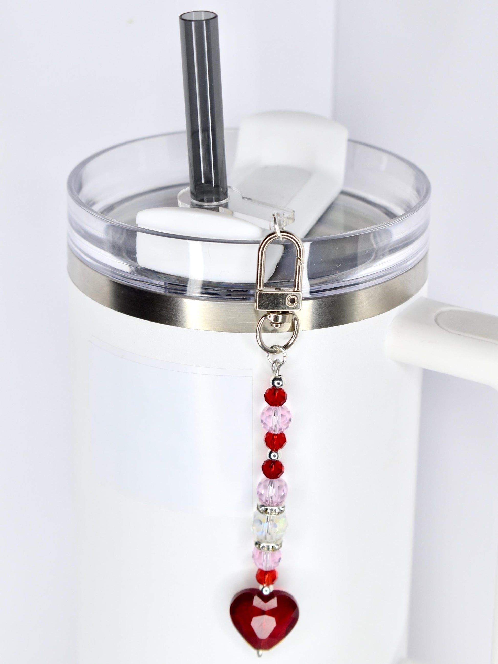 A white tumbler with a clear lid and a silver band. A beaded charm with red, pink, and clear beads and a red heart pendant hangs from the lid. A black straw is inserted into the lid.