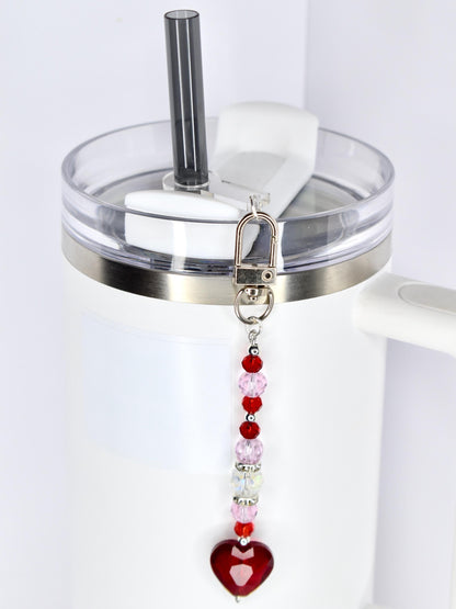 A white tumbler with a clear lid and a silver band. A beaded charm with red, pink, and clear beads and a red heart pendant hangs from the lid. A black straw is inserted into the lid.