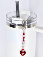 A white tumbler with a clear lid and a silver band. A beaded charm with red, pink, and clear beads and a red heart pendant hangs from the lid. A black straw is inserted into the lid.