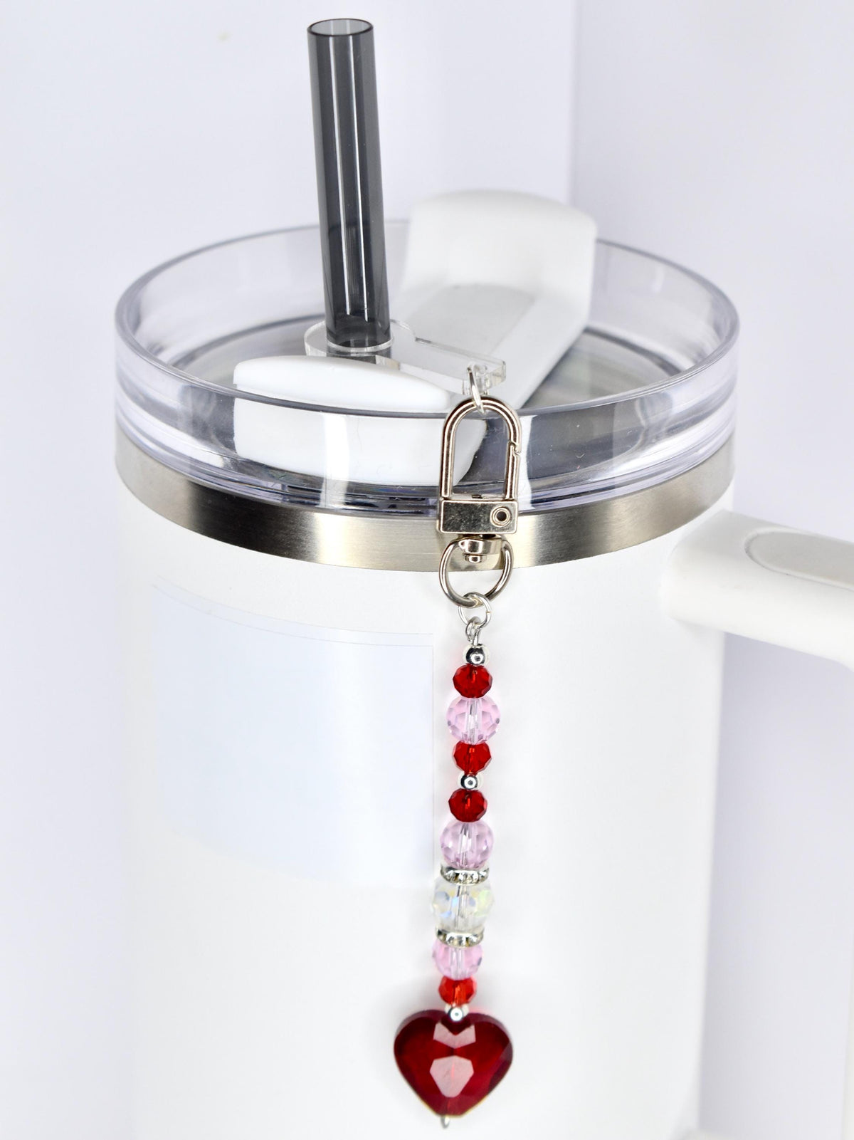 A white tumbler with a clear lid and a silver band. A beaded charm with red, pink, and clear beads and a red heart pendant hangs from the lid. A black straw is inserted into the lid.