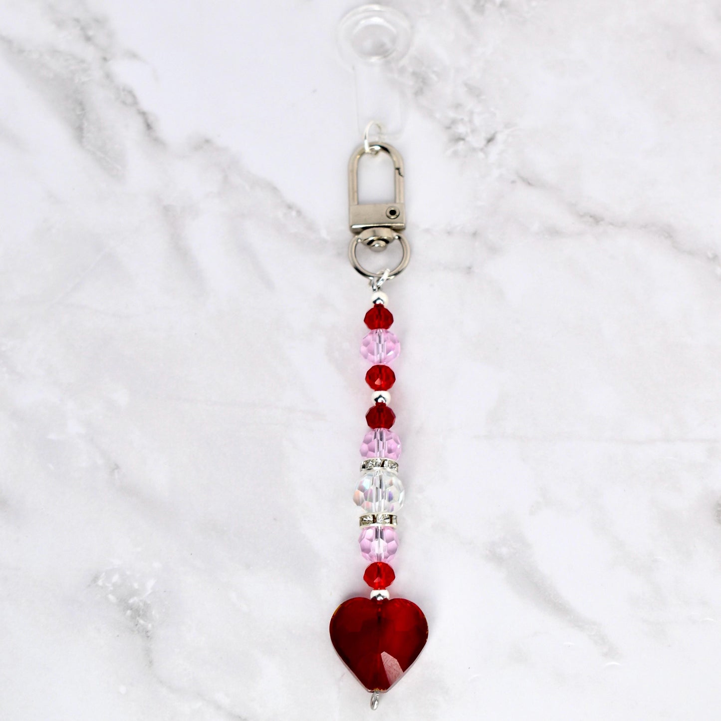 A beaded keychain with a silver clasp and a red heart charm. The keychain features a strand of pink, red, and clear beads. The heart charm is a deep red color.