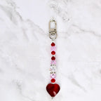 A beaded keychain with a silver clasp and a red heart charm. The keychain features a strand of pink, red, and clear beads. The heart charm is a deep red color.