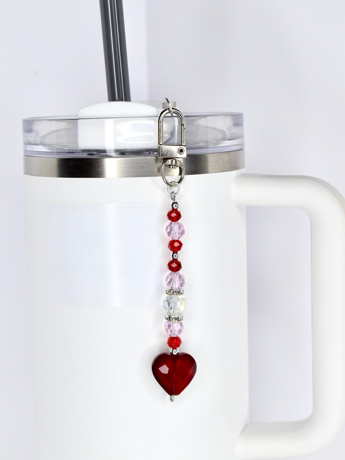 A beaded straw charm with a lobster clasp, featuring a heart-shaped red bead at the bottom. The charm has a mix of red, pink, and clear beads. It is attached to a white tumbler with a silver rim and a black straw.