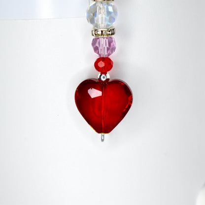 A red heart-shaped pendant with a silver bail, strung with a series of beads. The beads include clear, iridescent, pink, and red elements. The pendant is likely a piece of jewelry, such as a necklace charm or earring.