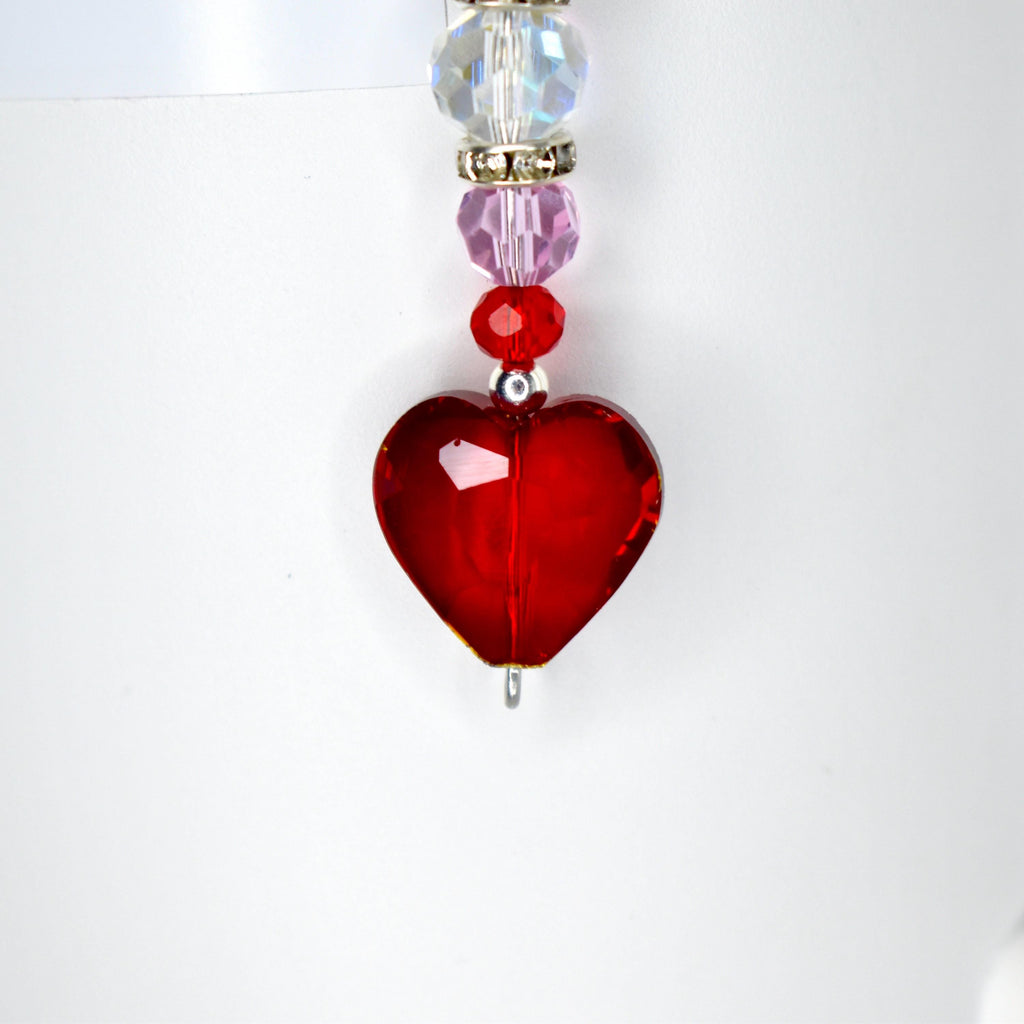 A red heart-shaped pendant with a silver bail, strung with a series of beads. The beads include clear, iridescent, pink, and red elements. The pendant is likely a piece of jewelry, such as a necklace charm or earring.
