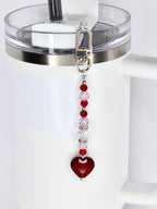 A beaded keychain with a silver clasp, featuring a strand of red, pink, and clear beads, culminating in a faceted, heart-shaped pendant. The keychain is attached to a white tumbler.